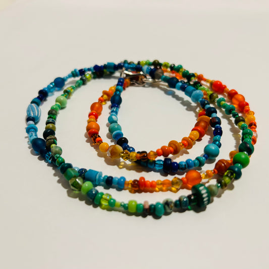 A long beaded necklace displayed laying on a surface, wound up to resemble bracelets. The necklace has 3 sections of various beads- orange, blue, & green. 
