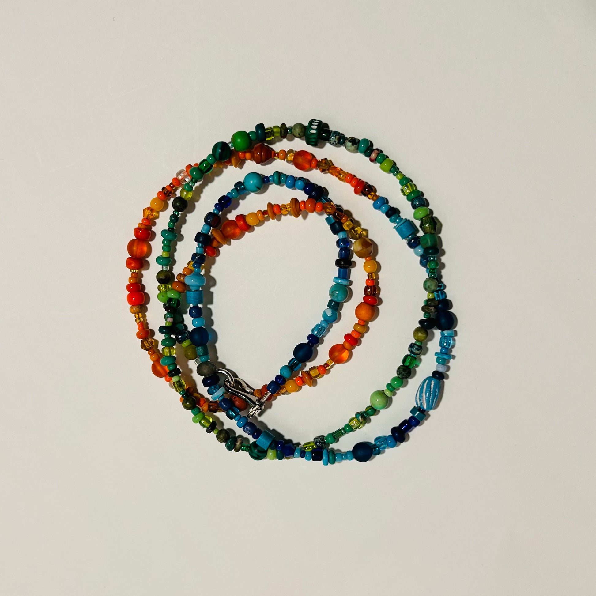 A long beaded necklace displayed laying on a surface, wound up to resemble bracelets. The necklace has 3 sections of various beads- orange, blue, & green. 