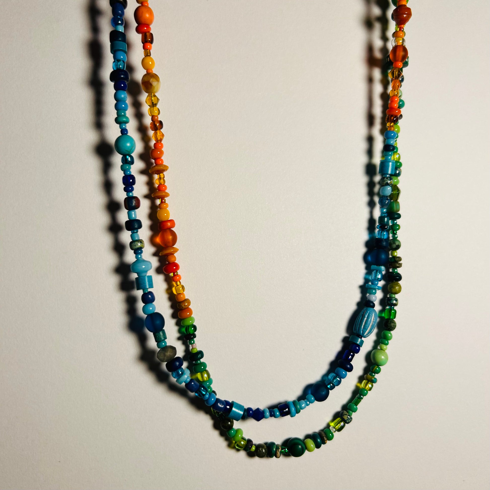 A long beaded necklace displayed hanging, doubled. The necklace has 3 sections of various beads- orange, blue, & green. Handmade beaded necklace, 37.5 inches in length with hook clasp. Made with a marvelous assortment of vintage & new beads from across the globe. Can be worn as a necklace, wrap choker, wrap bracelet, body chain, decorative belt.

