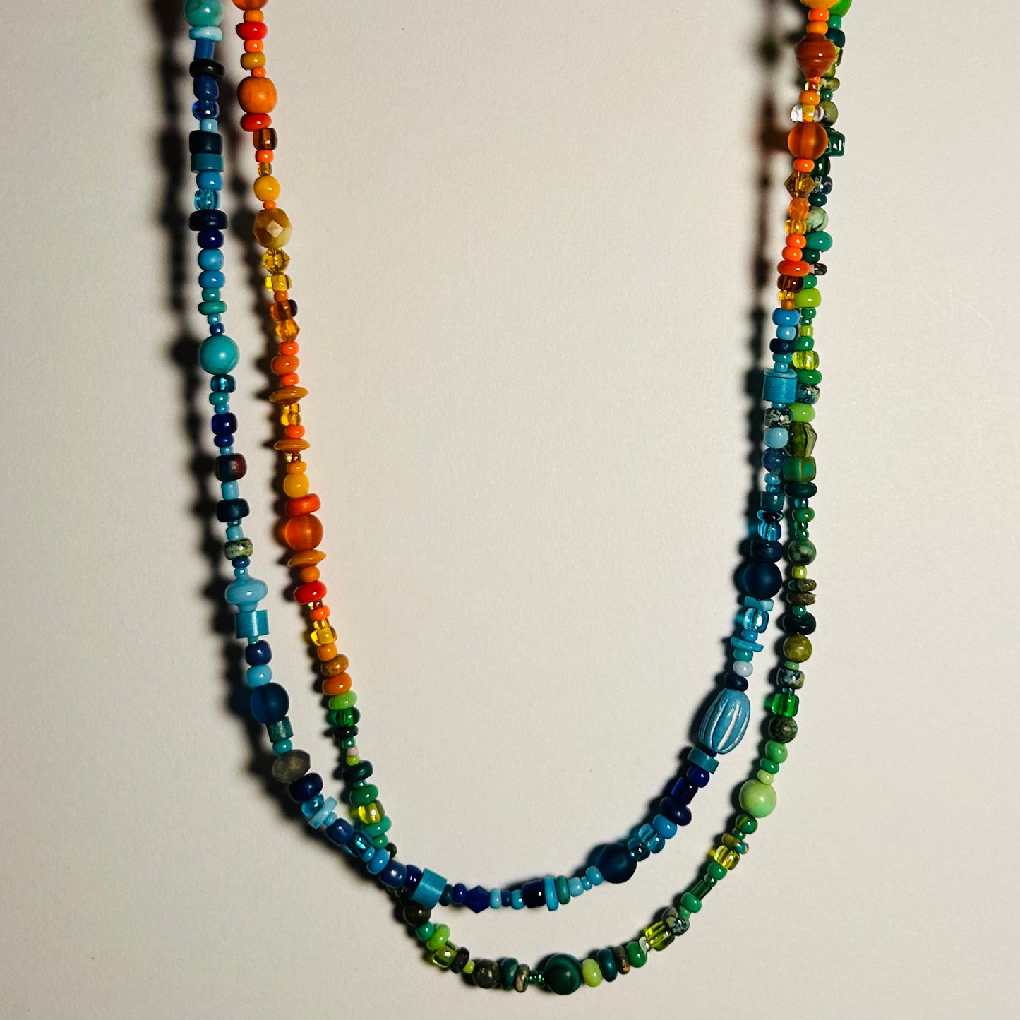A long necklace displayed hanging, doubled. The necklace has 3 sections of various beads- orange, blue, & green. Handmade beaded necklace, 37.5 inches in length with hook clasp. Made with a marvelous assortment of vintage & new beads from across the globe. Can be worn as a necklace, wrap choker, wrap bracelet, body chain, decorative belt.

